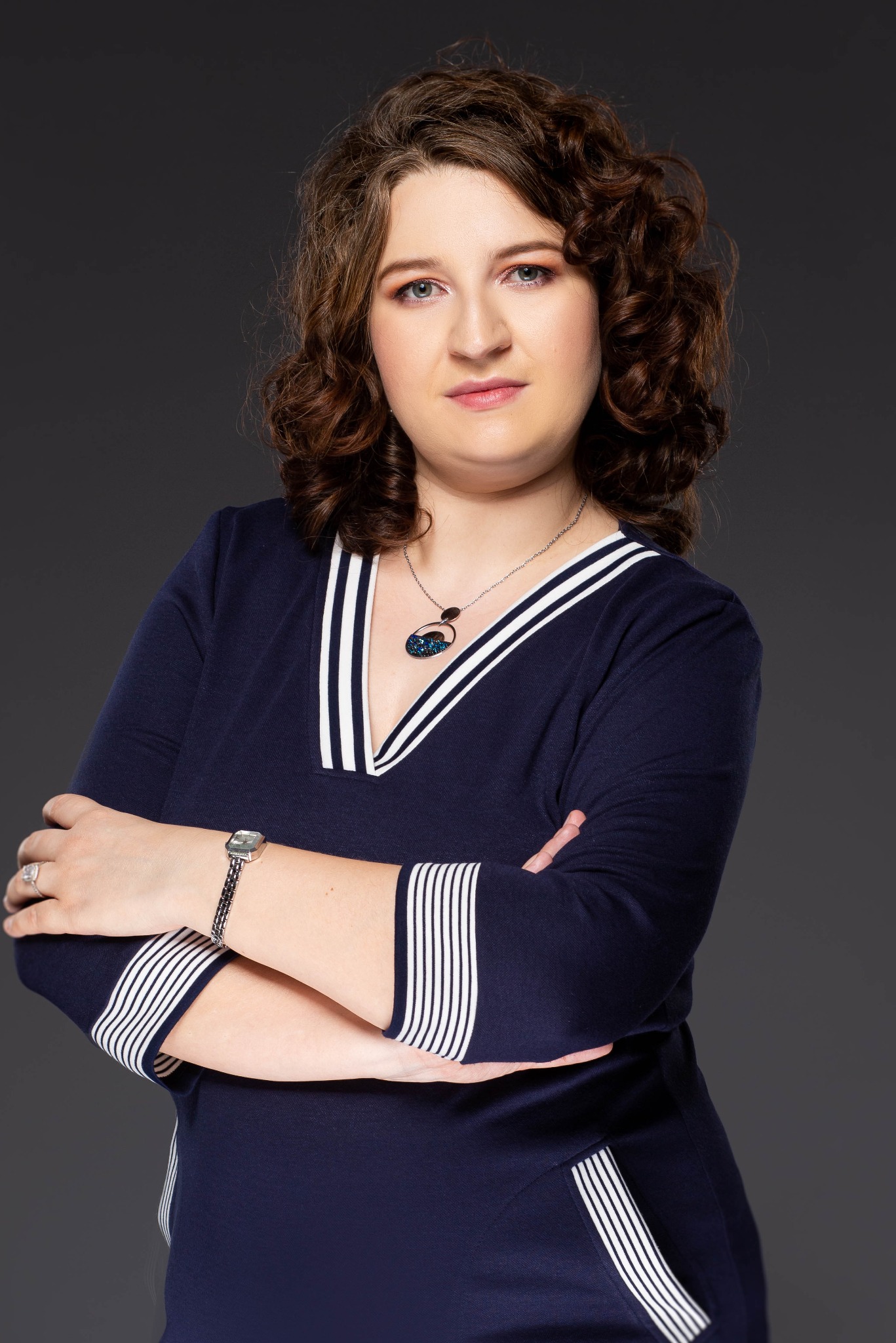 Portret kobiety z kręconymi włosami, ubranej w granatową sukienkę z białymi paskami, stojącej na ciemnoszarym tle z założonymi rękami, zegarkiem na nadgarstku i ozdobnym naszyjnikiem.