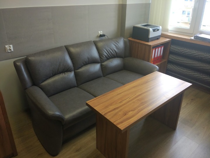 Kancelaria: Szary skórzany fotel i drewniany stolik kawowy w poczekalni, obok regał z segregatorami i drukarka przy oknie.