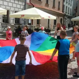 Grupa dzieci trzymająca dużą, kolorową chustę animacyjną na brukowanym placu miejskim, otoczonym budynkami i parasolami kawiarnianymi.