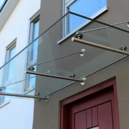 Szklany daszek nad wejściem do domu, wsparty na stalowych prętach i mocowaniach, widoczne detale mocowań i tekstura elewacji.