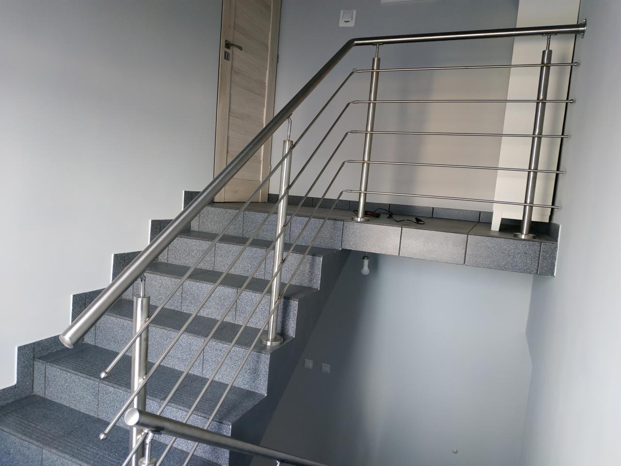 Schody wyłożone szarą płytką z zamontowaną metalową balustradą z poziomymi wypełnieniami, widok z dołu schodów.