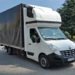 Biały samochód dostawczy Renault Master z plandeką na parkingu z kostki brukowej, widok z przodu, w tle budynki mieszkalne i zieleń.