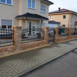 Czarne, kute ogrodzenie z bramką, osadzone na niskim murku z piaskowca, oddzielające posesję od chodnika z kostki brukowej.