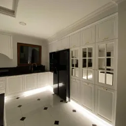 Elegancka, biała kuchnia na wymiar z czarnym blatem i złotymi akcentami, podświetlona od dołu, z czarną lodówką i oknem w drewnianej ramie.