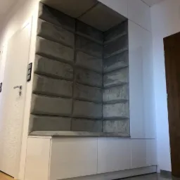 Wnęka z siedziskiem obita szarymi panelami tapicerowanymi w przedpokoju, białe szafki na górze i pod siedziskiem.