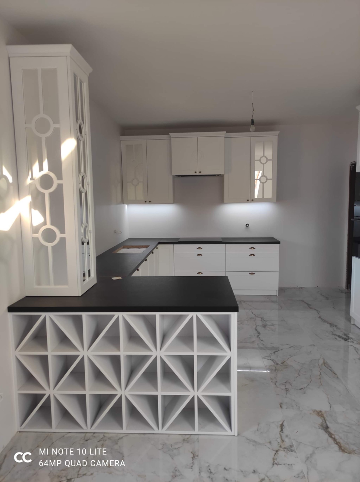 Nowoczesna biała kuchnia z czarnym blatem, szafkami z dekoracyjnymi przeszkleniami i regałem na wino w dolnej części wyspy kuchennej, na podłodze marmurowe płytki.