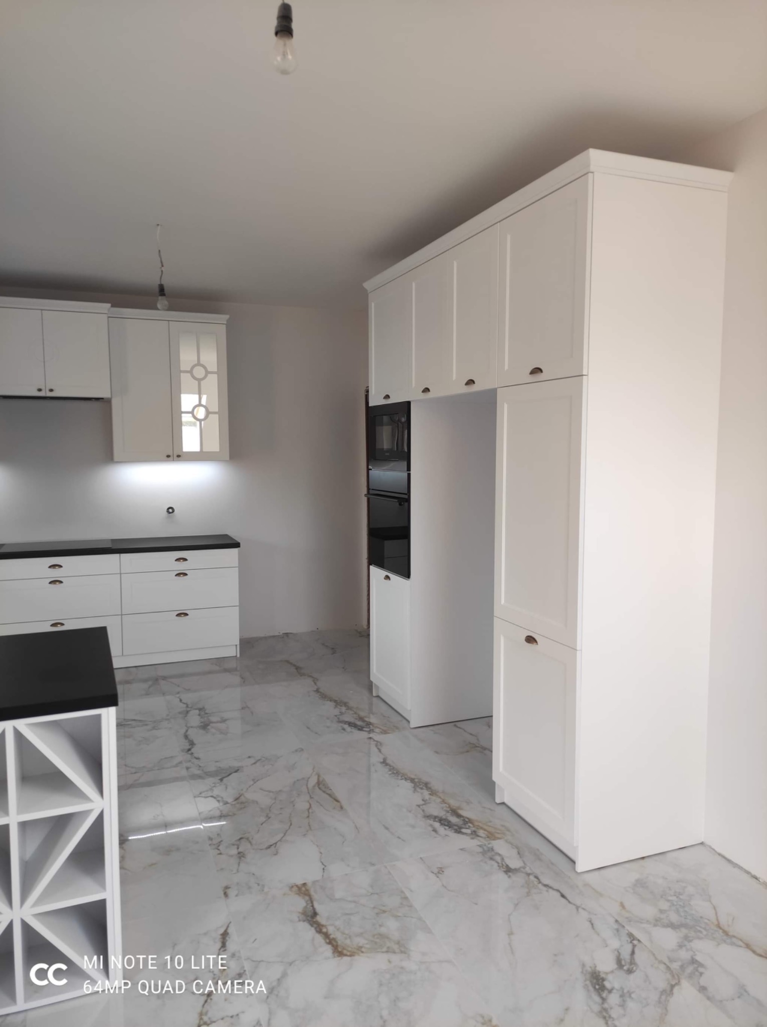 Wnętrze kuchni w trakcie wykańczania: białe szafki z ciemnymi blatami, marmurowe płytki podłogowe, widoczne niedokończone instalacje oświetleniowe.