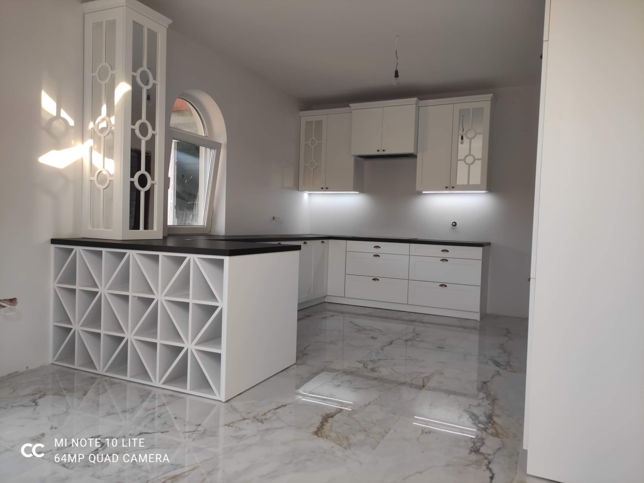 Biała, nowoczesna zabudowa kuchenna z czarnym blatem, dekoracyjnymi przeszkleniami w szafkach i ażurowym regałem na wino, na tle marmurowej podłogi.