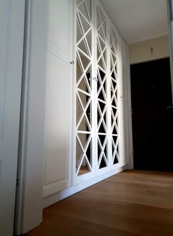 Biała szafa wnękowa z geometrycznym, krzyżowym wzorem na frontach, widoczna na tle drewnianej podłogi i czarnych drzwi.