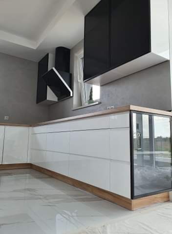 Nowoczesna kuchnia z białymi, lakierowanymi frontami, drewnianym blatem i czarnymi szafkami wiszącymi, widoczny fragment okna i marmurowa podłoga.