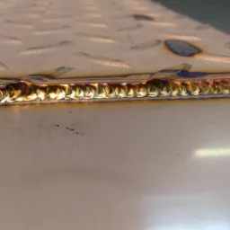 Szlif spawu na powierzchni metalowej z widoczną strukturą romboidalną, z efektem tęczy na krawędzi spoiny.