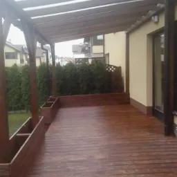 Zadaszony taras z drewnianą podłogą i drewnianą konstrukcją, z donicami wbudowanymi w balustradę, widok na ogród i sąsiednie domy.