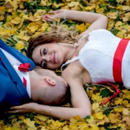 Para młoda leżąca na ziemi pokrytej żółtymi liśćmi, panna młoda w białej sukni z czerwoną szarfą, pan młody w niebieskim garniturze z czerwoną muszką.