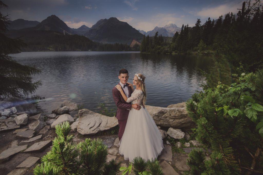Para młoda w objęciach na tle górskiego jeziora i szczytów, on w bordowym garniturze, ona w białej sukni z koronkowymi rękawami.