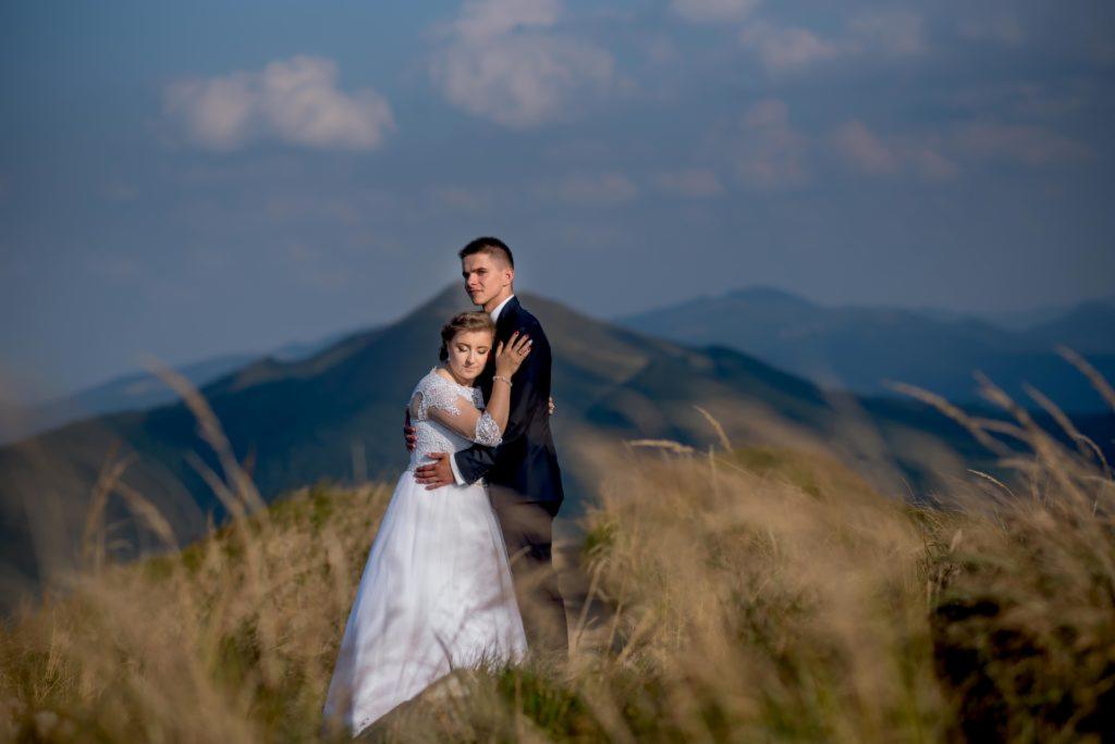 Para młoda w objęciach na tle gór i wysokiej trawy, panna młoda w sukni z koronkowymi rękawami, pan młody w ciemnym garniturze.
