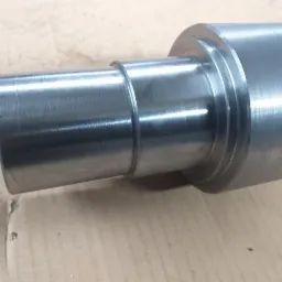 Szlifowany, metalowy wał z wyraźnymi śladami obróbki, widoczne stopnie na cylindrycznej powierzchni, leżący na kartonowym podłożu.