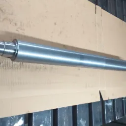 Szlifowany, metalowy wał z trzema pierścieniami na końcu, leżący na kartonie wewnątrz metalowej skrzyni.