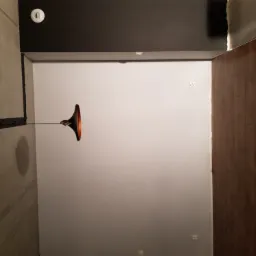 Wnętrze pokoju po remoncie: ściany pomalowane na jasny odcień szarości, ciemny sufit, drewniana okładzina ścienna po prawej stronie i nowoczesna lampa wisząca z miedzianym kloszem.