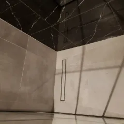 Wykończona kabina prysznicowa w Brzeźcach, z szarymi płytkami na ścianach i czarnymi płytkami imitującymi marmur na suficie, z odpływem liniowym w rogu.