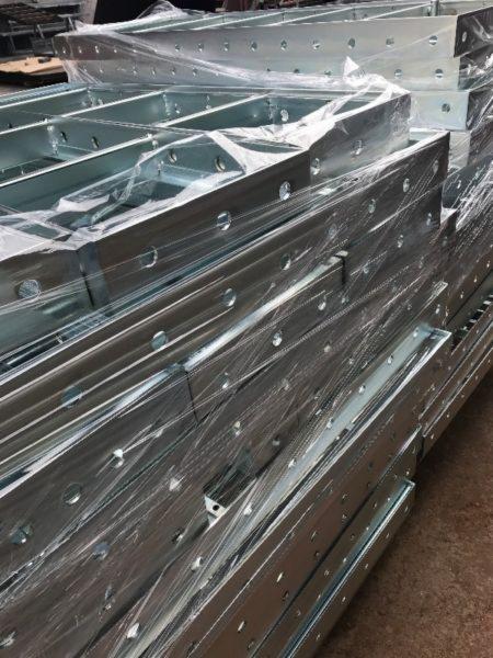 Stos ocynkowanych, metalowych belek konstrukcyjnych z otworami, owiniętych folią ochronną, przygotowanych do transportu lub montażu.