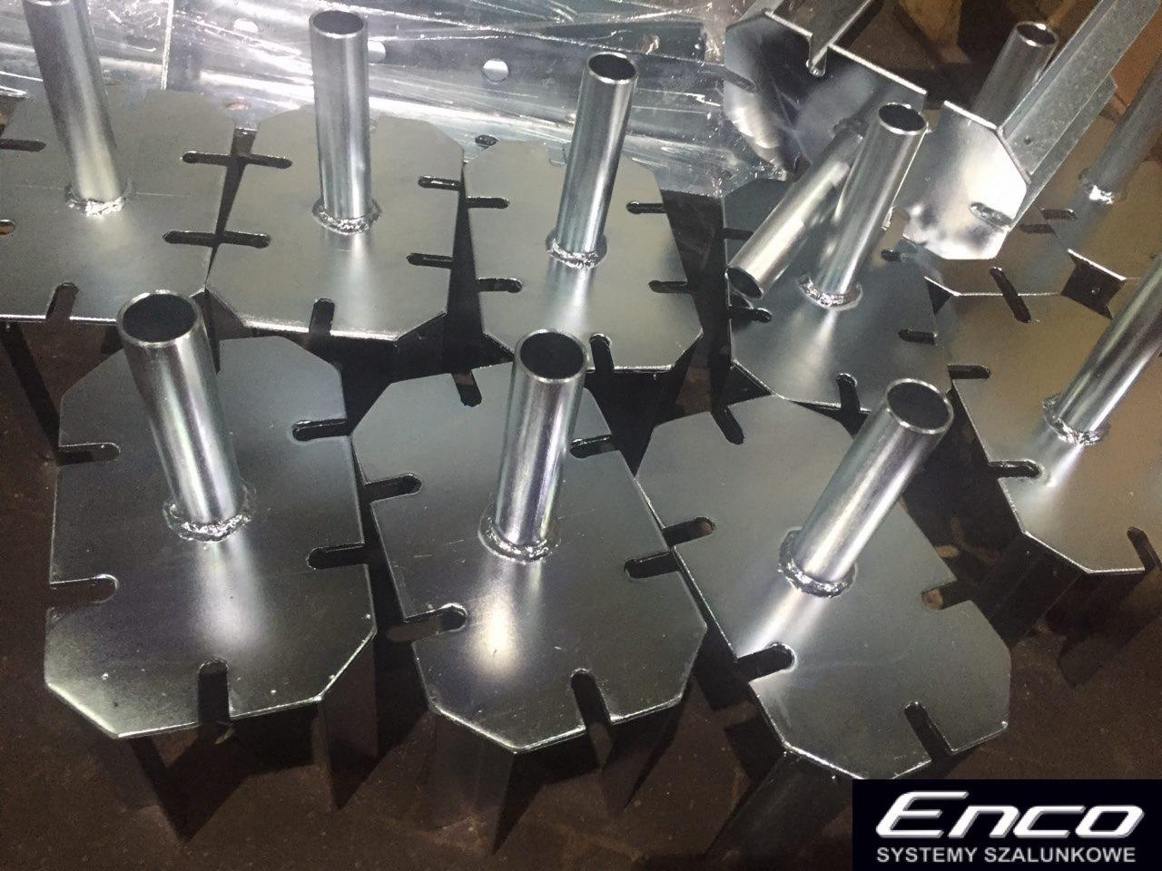 Metalowe, ocynkowane podstawki szalunkowe z przyspawanymi rurkami, ułożone obok siebie, widoczne spawy, w tle folia ochronna.