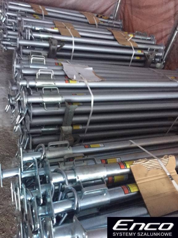 Stos ocynkowanych podpór budowlanych z uchwytami, zabezpieczonych kartonem i taśmą, w magazynie z widocznym logo producenta systemów szalunkowych.