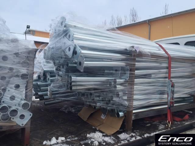 Stalowe, ocynkowane stemple budowlane, zabezpieczone folią stretch na palecie transportowej, widoczne mocowania i otwory, tło stanowią budynki magazynowe.