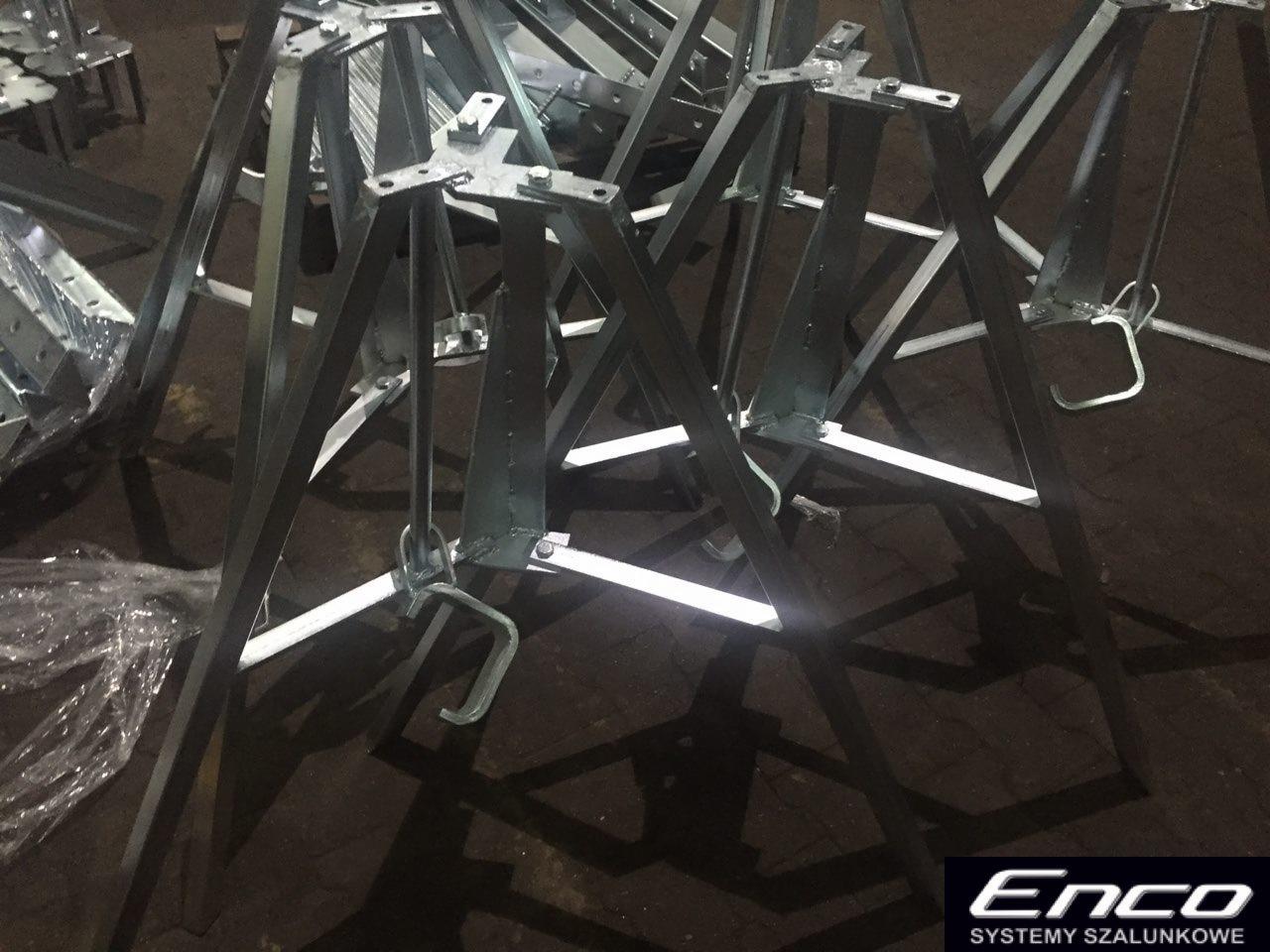 Stalowe stojaki szalunkowe z regulacją wysokości, widoczne ślady spawania i niedoskonałości malowania, logo producenta w prawym dolnym rogu.