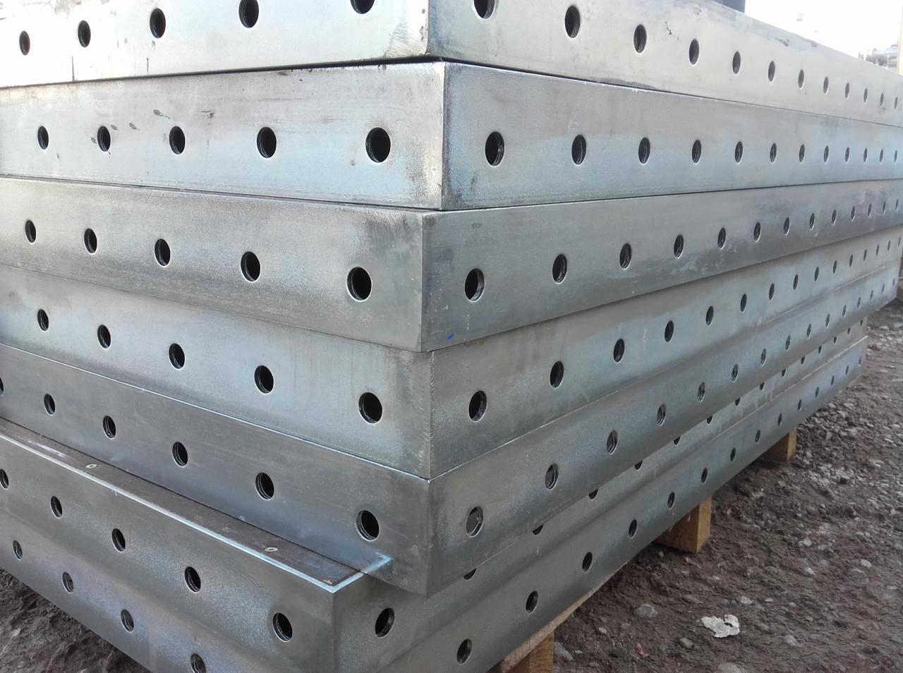 Stos ocynkowanych, metalowych szalunków budowlanych z regularnymi otworami, ułożonych na drewnianej palecie na zewnątrz.