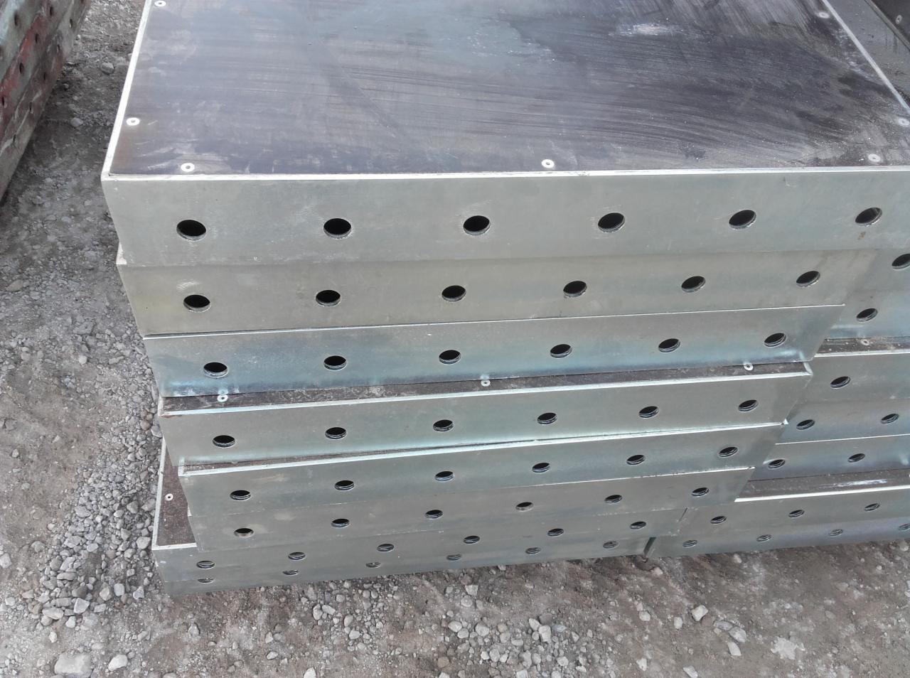 Stos szalunków budowlanych z metalową ramą i czarną płytą sklejkową, widoczne otwory montażowe, ułożone na żwirowym podłożu.