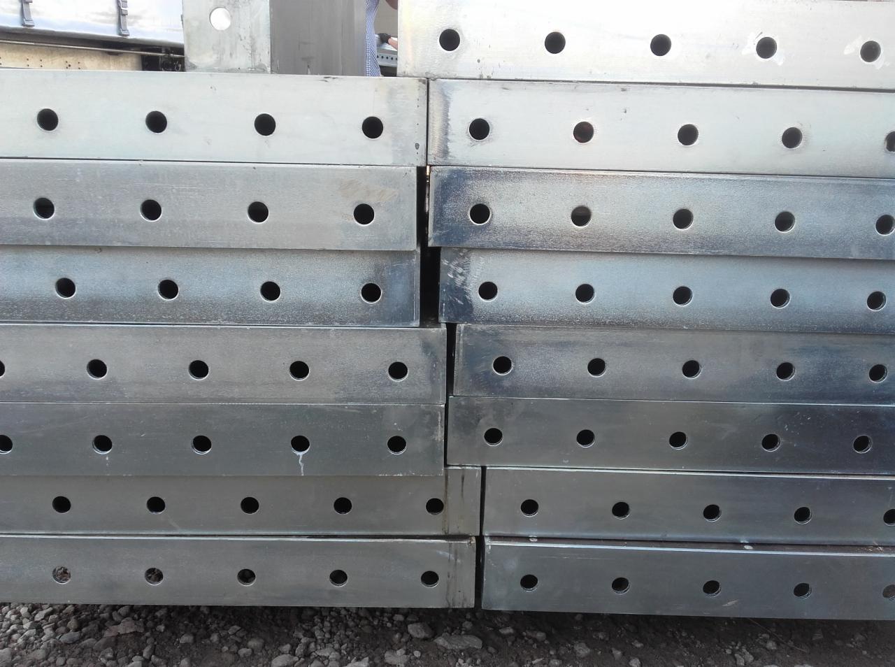Stos ocynkowanych, metalowych belek z otworami, prawdopodobnie do rusztowań budowlanych, ułożonych na ziemi.