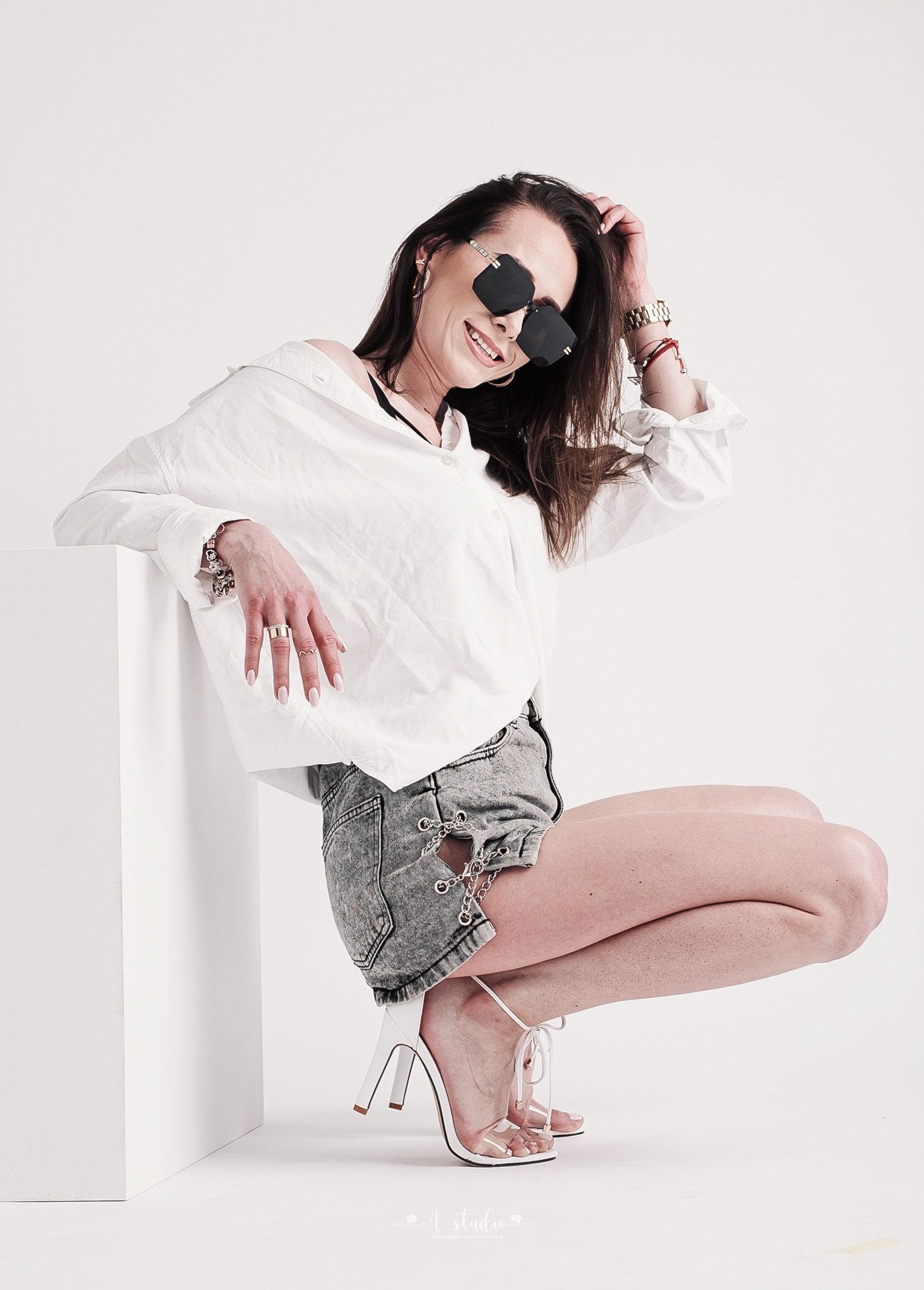 Uśmiechnięta kobieta w białej koszuli i dżinsowych szortach, pozująca w okularach przeciwsłonecznych i na wysokich obcasach, opierając się o biały sześcian w studiu.