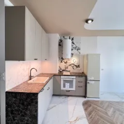 Nowoczesna kuchnia z marmurowymi blatami i podłogą, kremowymi szafkami, białym okapem i lodówką w stylu retro, oświetleniem LED pod górnymi szafkami i drewnianą podłogą w jodełkę na granicy...