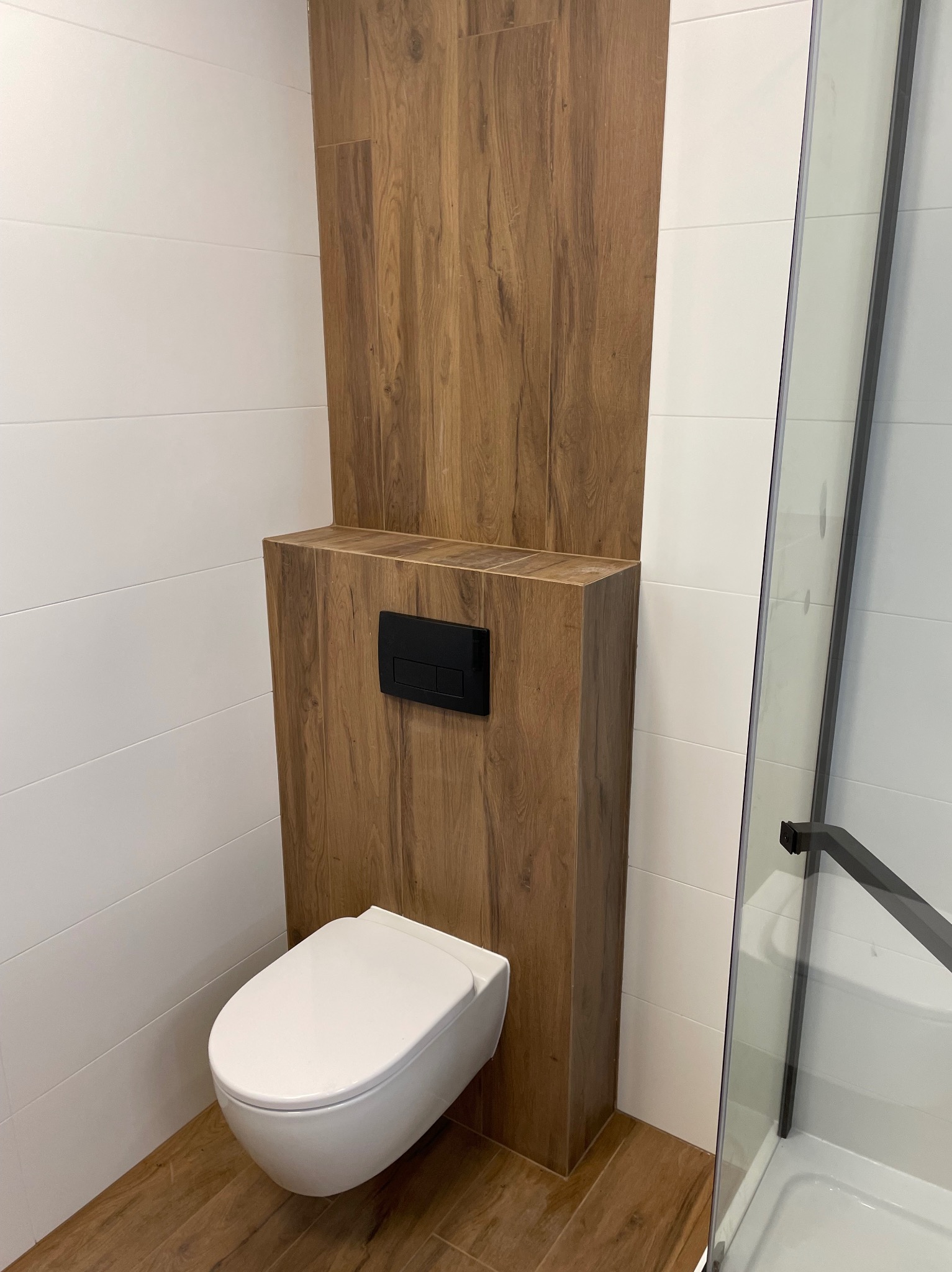 Nowoczesna łazienka po remoncie: podwieszana toaleta z drewnopodobną obudową i czarnym przyciskiem, białe, poziome płytki na ścianach, kabina prysznicowa z czarnymi ramami.