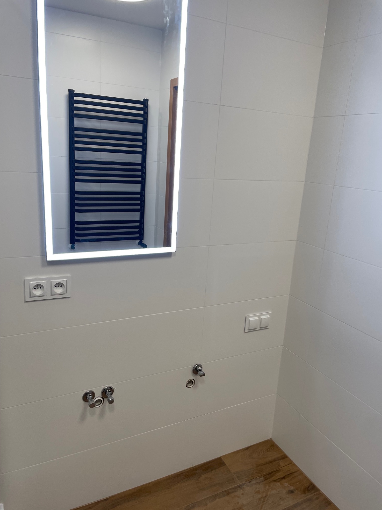 Biała łazienka z drewnopodobną podłogą. Na ścianie lustro z podświetleniem LED, w którym odbija się czarny grzejnik drabinkowy. Widoczne gniazdka i włączniki.