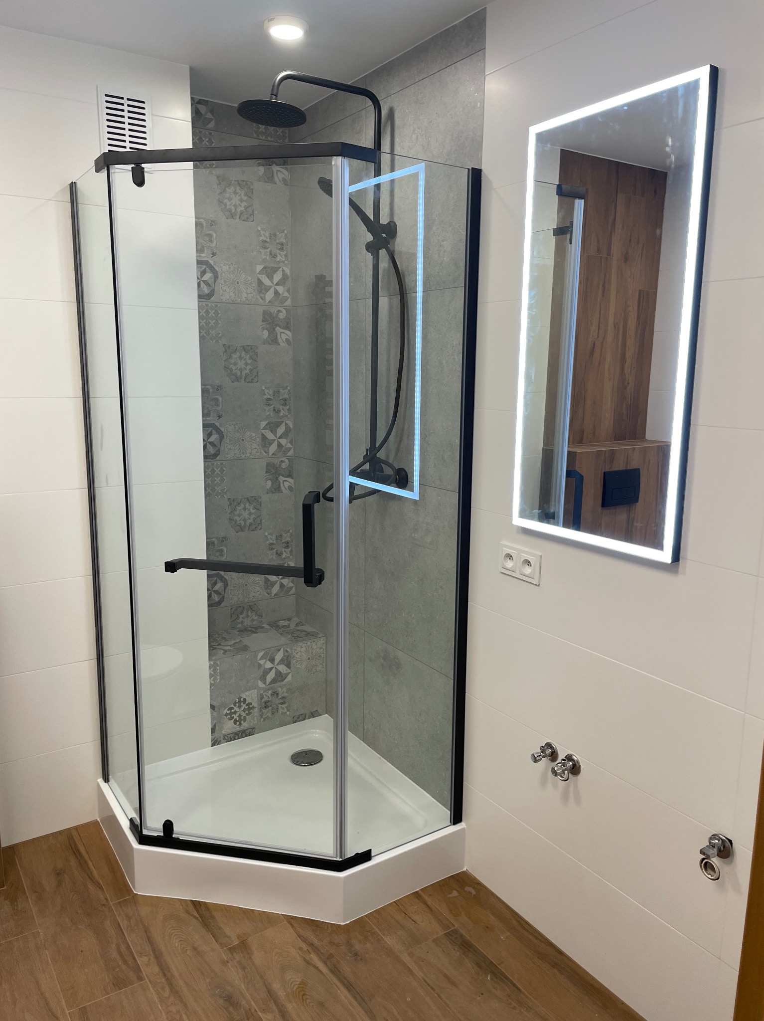 Nowoczesna kabina prysznicowa z czarnymi profilami i deszczownicą, obok podświetlane lustro. Białe płytki i drewnopodobna podłoga tworzą spójną całość.