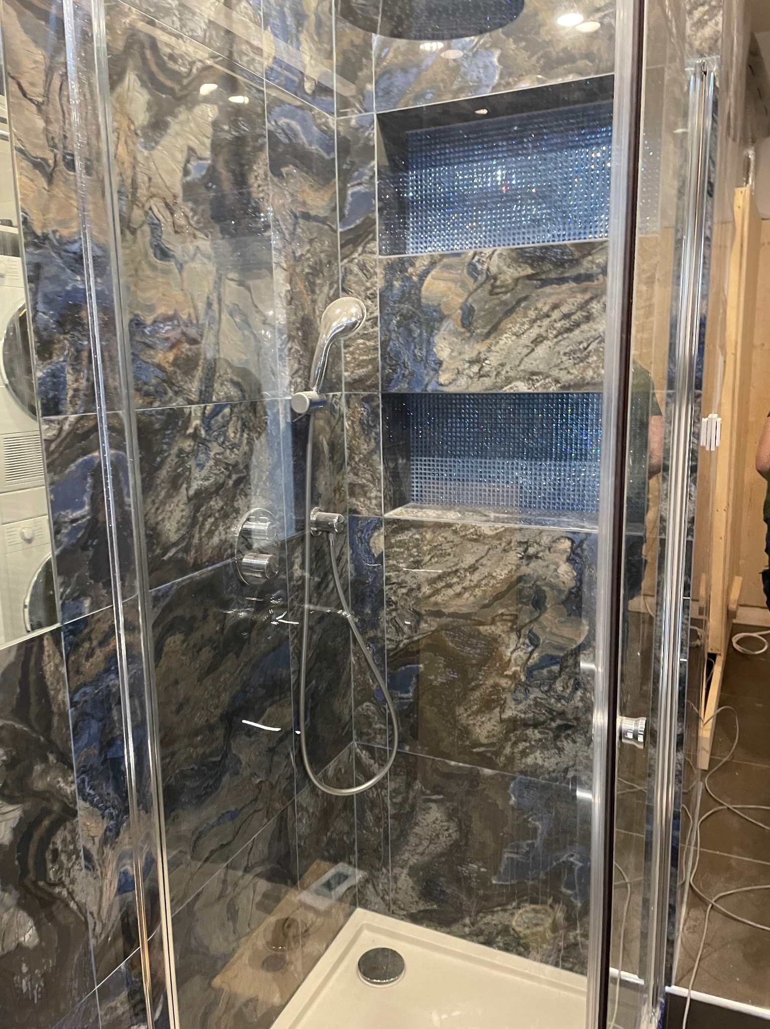 Kabina prysznicowa z ciemnymi, marmurkowymi płytkami i wnękami wyłożonymi mozaiką. Widoczna armatura i brodzik. Częściowo widoczna pralka i elementy instalacji.