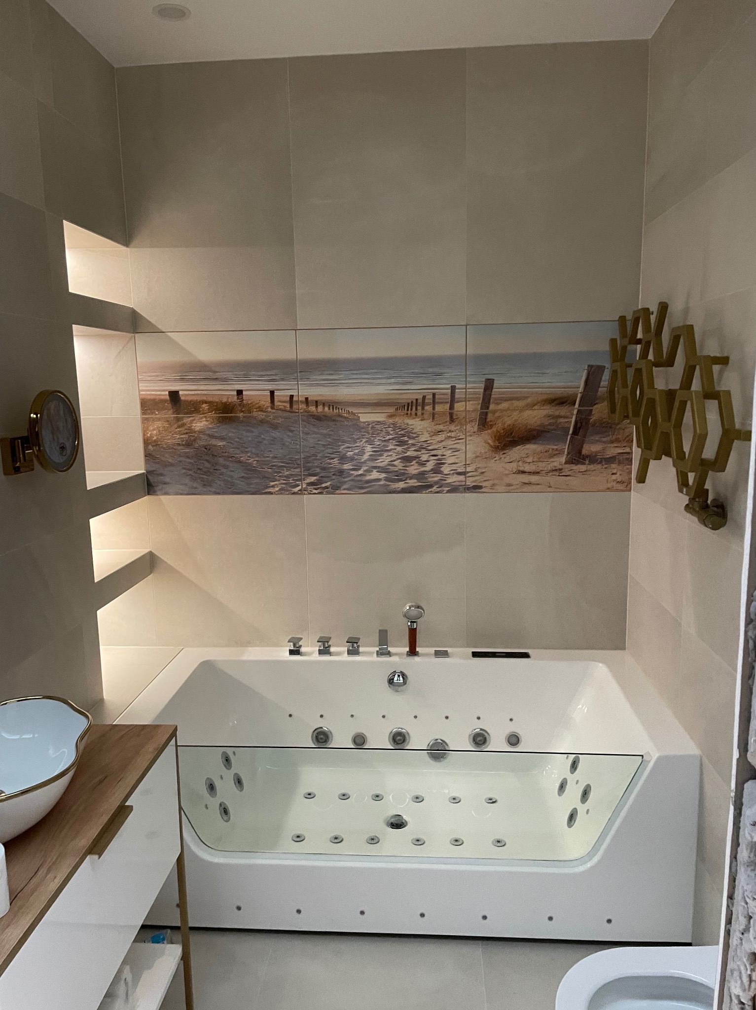 Nowoczesna łazienka z wanną z hydromasażem, wykończona płytkami z motywem plaży. Umywalka na drewnianym blacie i dekoracyjna złota ozdoba na ścianie.