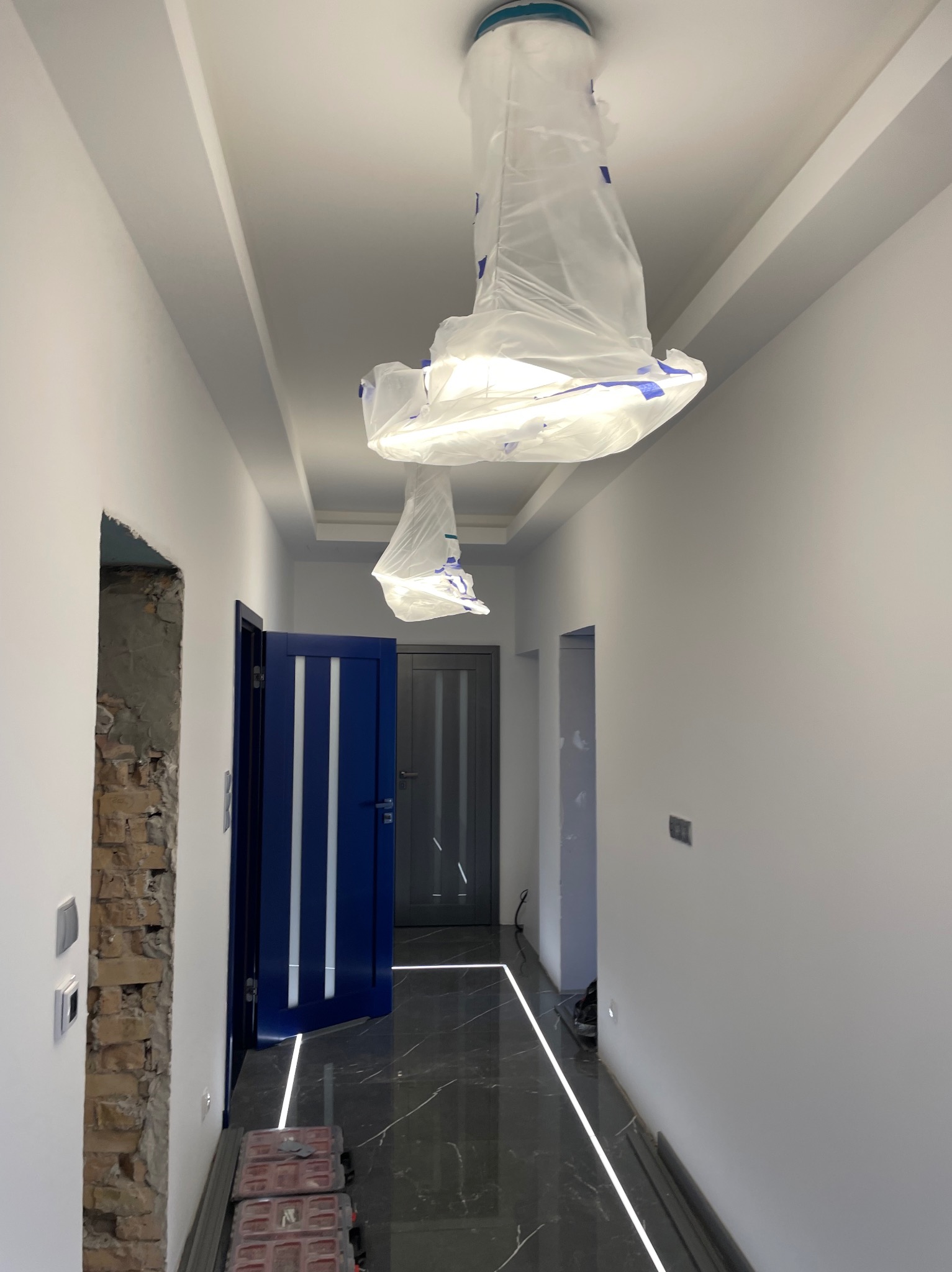 Korytarz w trakcie wykańczania: marmurowa podłoga z oświetleniem LED, pomalowane ściany, zabezpieczone lampy i otwarte drzwi w kolorze granatowym.