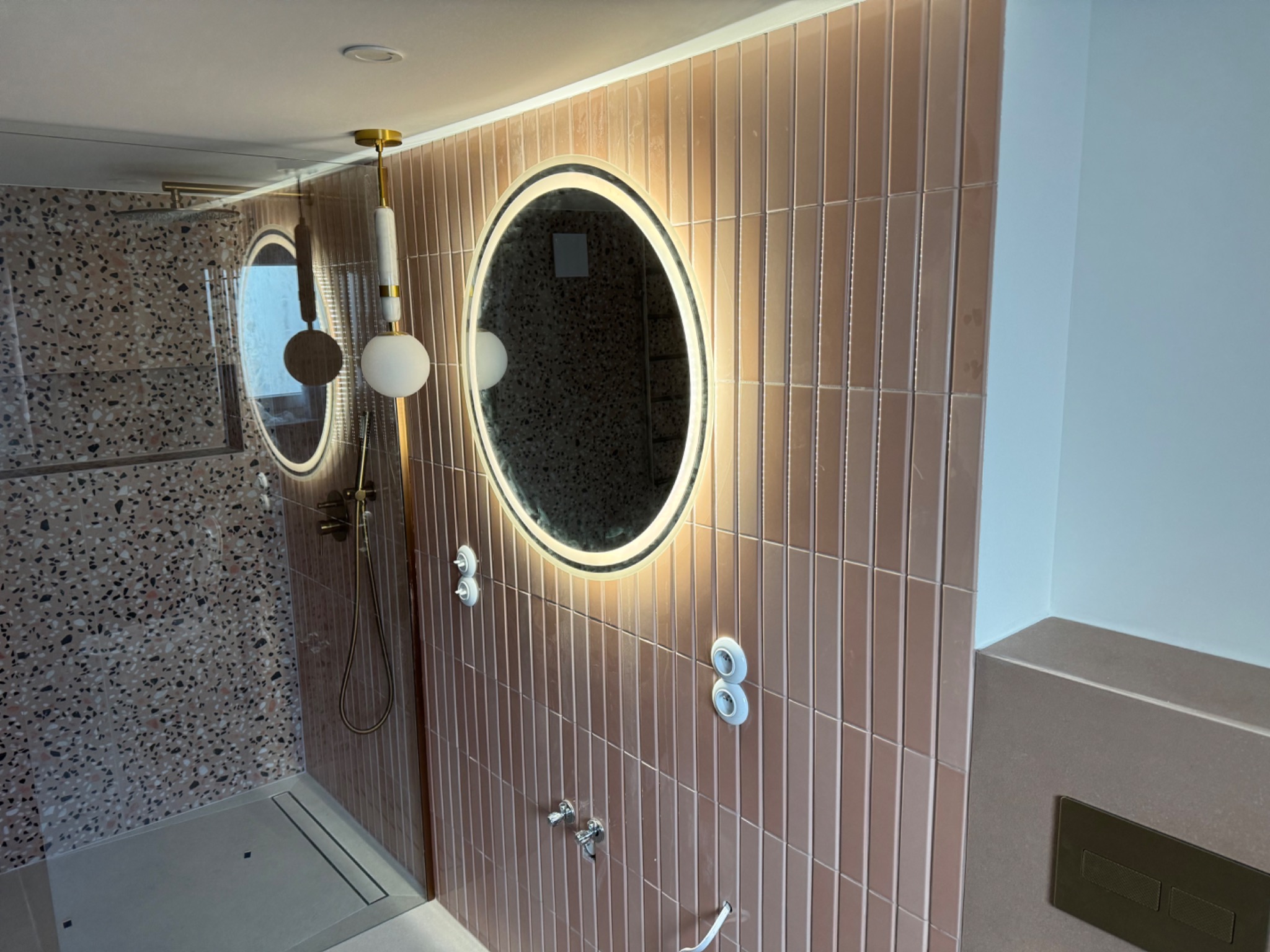 Nowoczesna łazienka po remoncie: różowe, pionowo ułożone płytki, okrągłe lustro z podświetleniem LED, armatura w kolorze srebra i kabina prysznicowa typu walk-in.