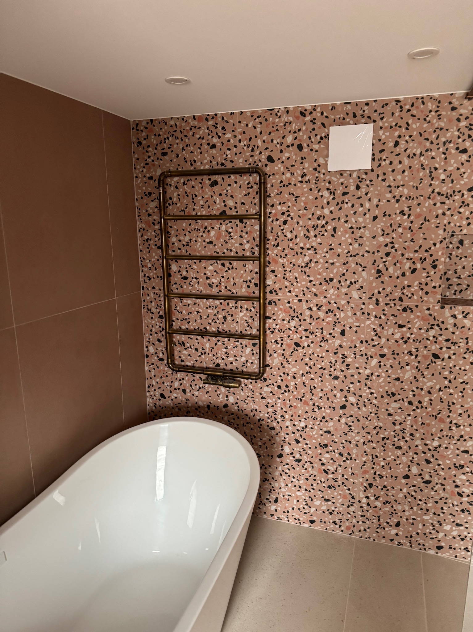 Nowoczesna łazienka z wanną wolnostojącą, grzejnikiem drabinkowym w kolorze mosiądzu i ścianą wykończoną płytkami imitującymi lastryko w odcieniach różu i czerni.