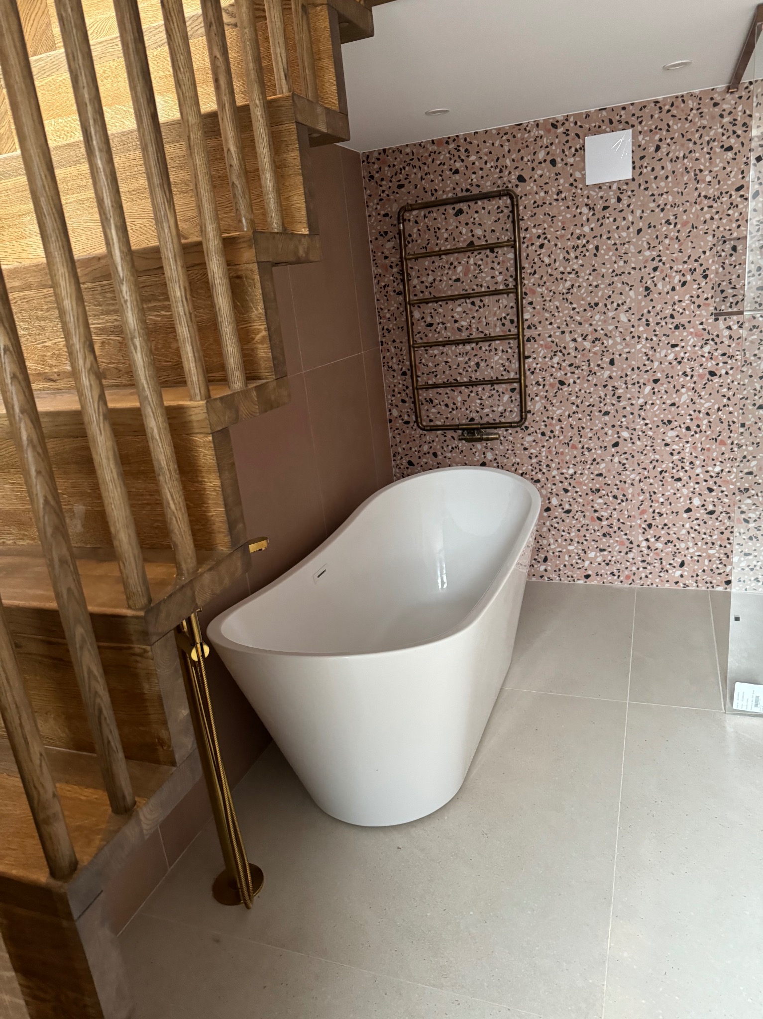 Nowoczesna łazienka z wolnostojącą białą wanną, złotą armaturą i grzejnikiem drabinkowym na ścianie wyłożonej mozaiką w odcieniach różu i czerni. Drewniane schody w tle.