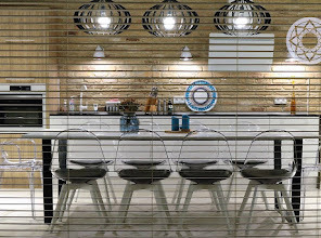 Nowoczesna kuchnia z ceglaną ścianą, oświetleniem wiszącym w stylu industrialnym i przezroczystymi krzesłami przy białym stole. Widoczne elementy dekoracyjne na ścianie i blacie.