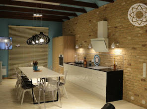 Nowoczesna kuchnia z ceglaną ścianą, białymi szafkami i blatem w kolorze czarnym, oświetlona wiszącymi lampami z dekoracyjnymi kloszami. Nad blatem rząd halogenowych punktowych świateł. Na ścianie...