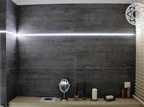 Nowoczesna łazienka z szarymi płytkami imitującymi beton, podświetleniem LED wzdłuż ściany, lustrem kosmetycznym, świecami i roletami w oknie.