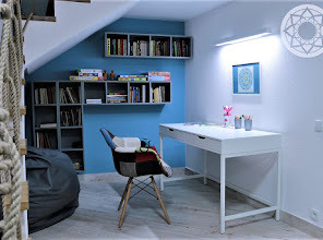 Aranżacja kącika do pracy pod schodami: białe biurko z szufladami, patchworkowe krzesło, półki z książkami na błękitnej ścianie, marmurowa podłoga, oświetlenie LED.