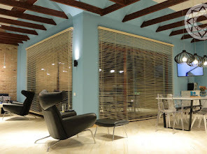 Jasne wnętrze z drewnianymi belkami stropowymi i dużymi oknami zasłoniętymi poziomymi żaluzjami. Niebieskie ściany, nowoczesne czarne fotele i przezroczyste krzesła przy stole. Uwagę przyciąga...