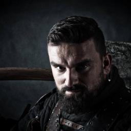 Kodama Foto - Portret brodatego mężczyzny w skórzanej zbroi, trzymającego topór na ramieniu, z intensywnym spojrzeniem w obiektyw.