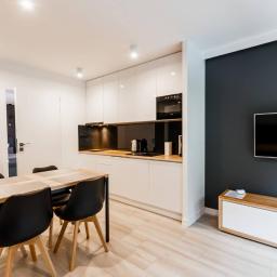 Kodama Foto - Jasne wnętrze mieszkania z aneksem kuchennym, drewnianym stołem z czarnymi krzesłami, białą zabudową kuchenną z czarnym panelem szklanym i drewnianym blatem, ciemnoszarą ścianą z telewizorem...
