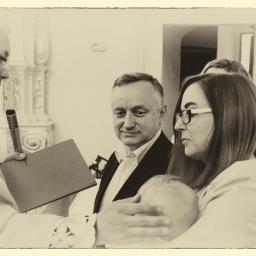 Kodama Foto - Ksiądz z mikrofonem prowadzi ceremonię, rodzice trzymają dziecko podczas chrztu, stonowana kolorystyka.